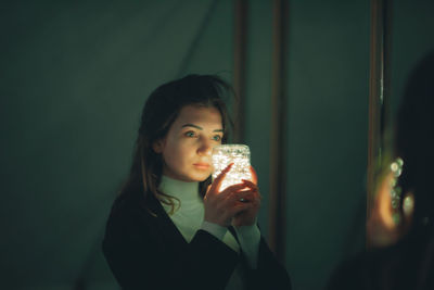 Young woman holding illuminated lighting equipment reflecting on mirror in darkroom