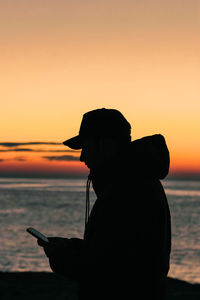 Silhouette man using smart phone by sea against sky during sunset