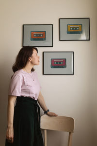 Adult female leaning on chair and admiring framed images of cassettes hanging on wall at home