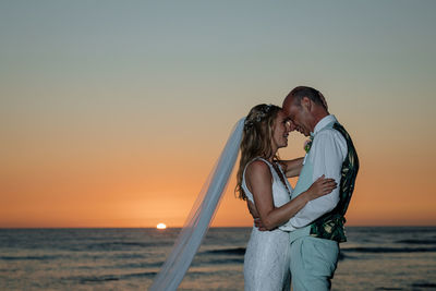 Couple standing by sea against sky during sunset