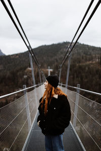 Female tourist in warm clothes walking along metal suspension bridge in highlands and enjoying vacation in autumn