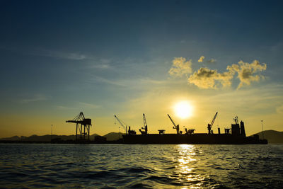 Silhouette cranes at harbor against sky during sunset