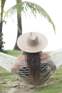 Rear view of woman wearing hat while relaxing on hammock over field