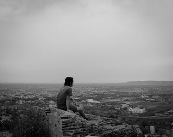 Man looking at cityscape while sitting on retaining wall against sky