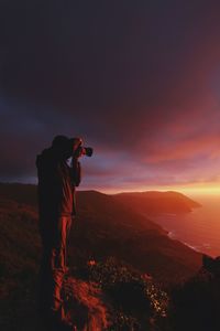 Man photographing on rock against sky during sunset