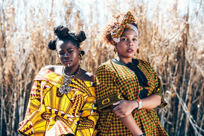 Self assured black female models in stylish clothes with authentic african prints looking at camera while standing together against dry grass in countryside