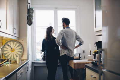 Rear view of couple talking while standing by dining table against window at home