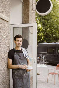 Male cafe owner wearing apron holding coffee cup leaning on wall