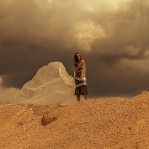 Low angle view of woman wrapped in plastic standing on sand dune against cloudy sky