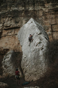 Rear view of man holding rope while friend climbing rock in forest