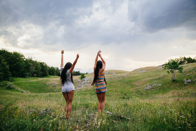 Rear view of women with arms raised standing on field against sky