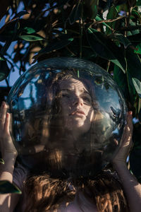 Close-up of young woman wearing glass ball on head against leaves