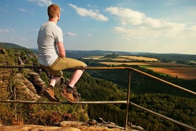 Short hair man sit on handrail at peak of rock and watch to land. hiker with grey shirt and pants