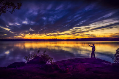 Man photographing lake while standing on rock against dramatic sky during sunset
