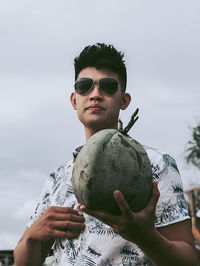 Portrait of young man holding sunglasses against sky