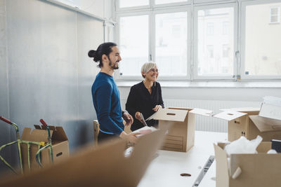 Smiling young businessman with female colleague unpacking cardboard boxes at table in new office