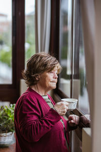 Side view of thoughtful senior lady with cup of hot drink looking out window and contemplating in cozy room at home