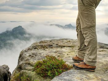 Long tired male legs in hiking trousers stay for moment on peak of rock above valley.