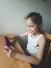 Portrait of smiling girl using mobile phone on table against wall at home