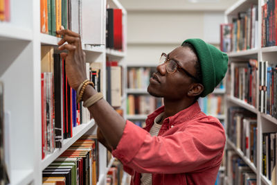 Puzzled concentrated black man student choosing need literature for study on bookshelves in library