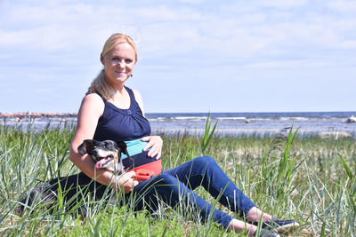 Portrait of smiling pregnant woman with dog sitting on grass against sky