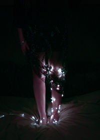 Low section of woman with illuminated string lights standing on bed in darkroom