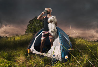 Man having drink while holding cat in tent on grassy field