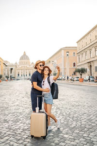 Happy young couple of travellers with hats and a suitcase hugging in the city and enjoying romance. 