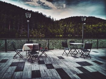 Empty chairs and table by lake against sky