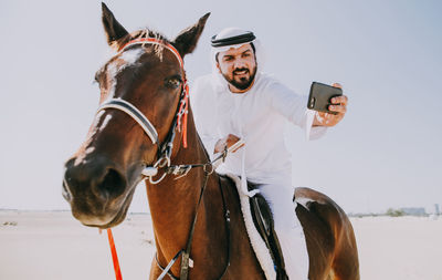 Man using mobile phone while horseback riding against sky