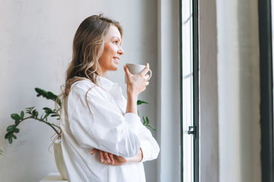 Young smiling blonde woman with long hair in white shirt rest and drinking tea near window in office