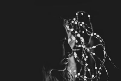 Woman looking away by illuminated string light against black background
