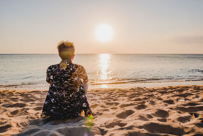 Rear view of man sitting on beach against sky during sunset