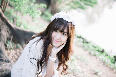 Portrait of beautiful young woman wearing white dress and wreath sitting on tree root in forest