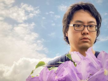 Portrait of young man behind purple clematis flowers against cloudy blue sky.