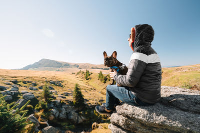 Young man with dog sitting on rock against sky