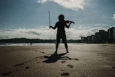 Full length of man playing violin standing on beach against sky