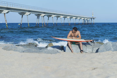 Shirtless man holding surfboard while sitting on rock at beach