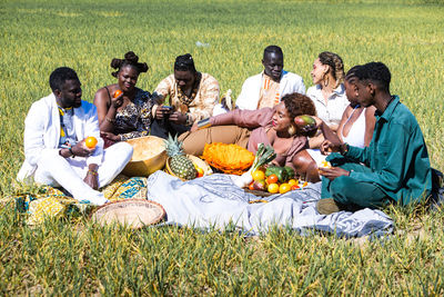 Ground level of group of stylish black woman with banana smiling with closed eyes and raising arm while lying on blanket amidst friends during picnic in grassy field on sunny summer day