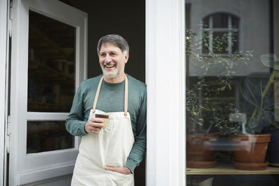 Smiling mature male owner looking away while having coffee at doorway of art studio