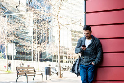 Front view of a bearded man using phone leaning on office building wall