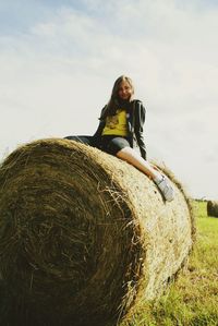 Young woman sitting on hay bale at field against sky during sunny day