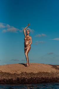 A pregnant girl in gold glitter stands on a coral stone in the middle of the ocean