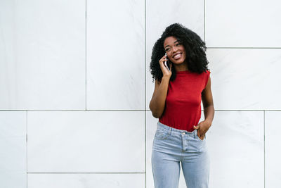 Smiling young ethnic woman in casual outfit standing near white tiled wall and holding hand in pocket while speaking on cellphone and looking away