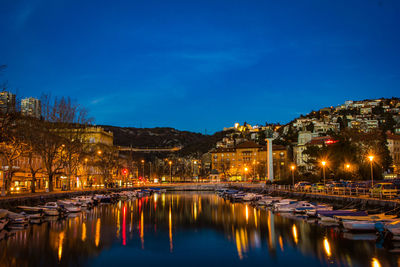 Illuminated buildings by river against blue sky at night