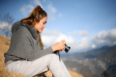 Side view young woman sitting on rock with camera against sky