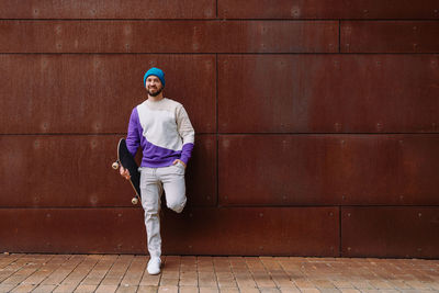 Young man with skateboard standing against wall