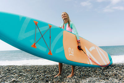 A young woman carries a sup board to the sea and smiles.