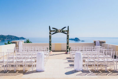 Empty chairs arranged on shore against clear blue sky during wedding ceremony