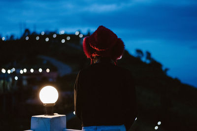 Rear view of woman standing by illuminated lamp at dusk